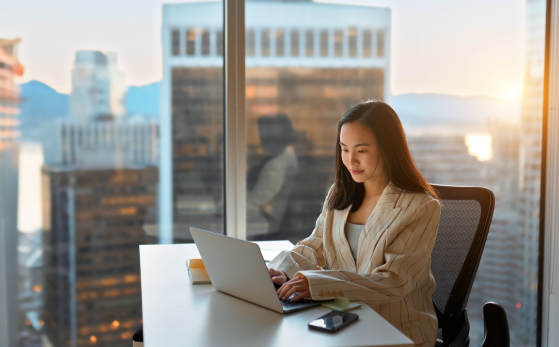 Young busy Asian business woman executive working on laptop in corporate office. Professional businesswoman marketing sales manager using computer technology sitting at table, city view from window.